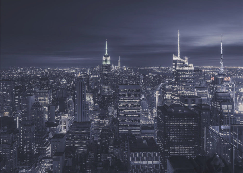 A dramatic nighttime photograph of the New York City skyline, viewed from a high vantage point. The cityscape is a sea of illuminated skyscrapers under a dark, moody sky, with the entire image cast in a cool blue and white monochrome tone and presented in a silver frame. Artwork