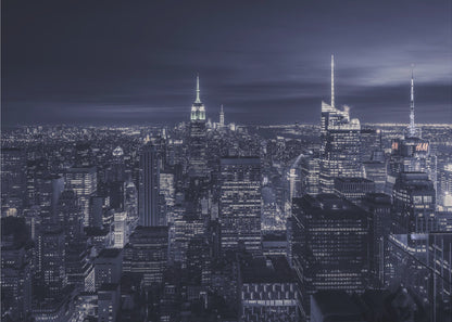 A dramatic nighttime photograph of the New York City skyline, viewed from a high vantage point. The cityscape is a sea of illuminated skyscrapers under a dark, moody sky, with the entire image cast in a cool blue and white monochrome tone and presented in a silver frame. Artwork