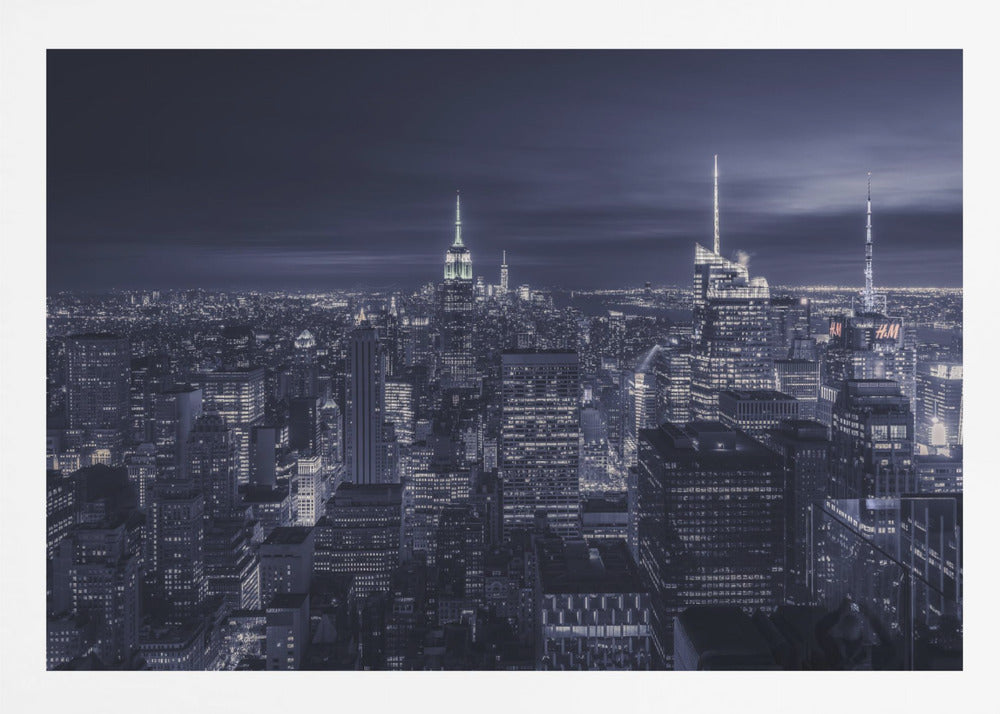 A dramatic nighttime photograph of the New York City skyline, viewed from a high vantage point. The cityscape is a sea of illuminated skyscrapers under a dark, moody sky, with the entire image cast in a cool blue and white monochrome tone and presented in a silver frame. Artwork