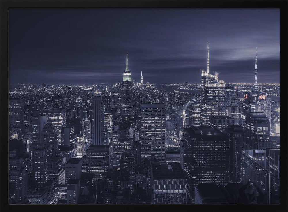 A dramatic nighttime photograph of the New York City skyline, viewed from a high vantage point. The cityscape is a sea of illuminated skyscrapers under a dark, moody sky, with the entire image cast in a cool blue and white monochrome tone and presented in a silver frame. Artwork
