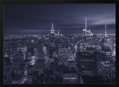 A dramatic nighttime photograph of the New York City skyline, viewed from a high vantage point. The cityscape is a sea of illuminated skyscrapers under a dark, moody sky, with the entire image cast in a cool blue and white monochrome tone and presented in a silver frame. Artwork