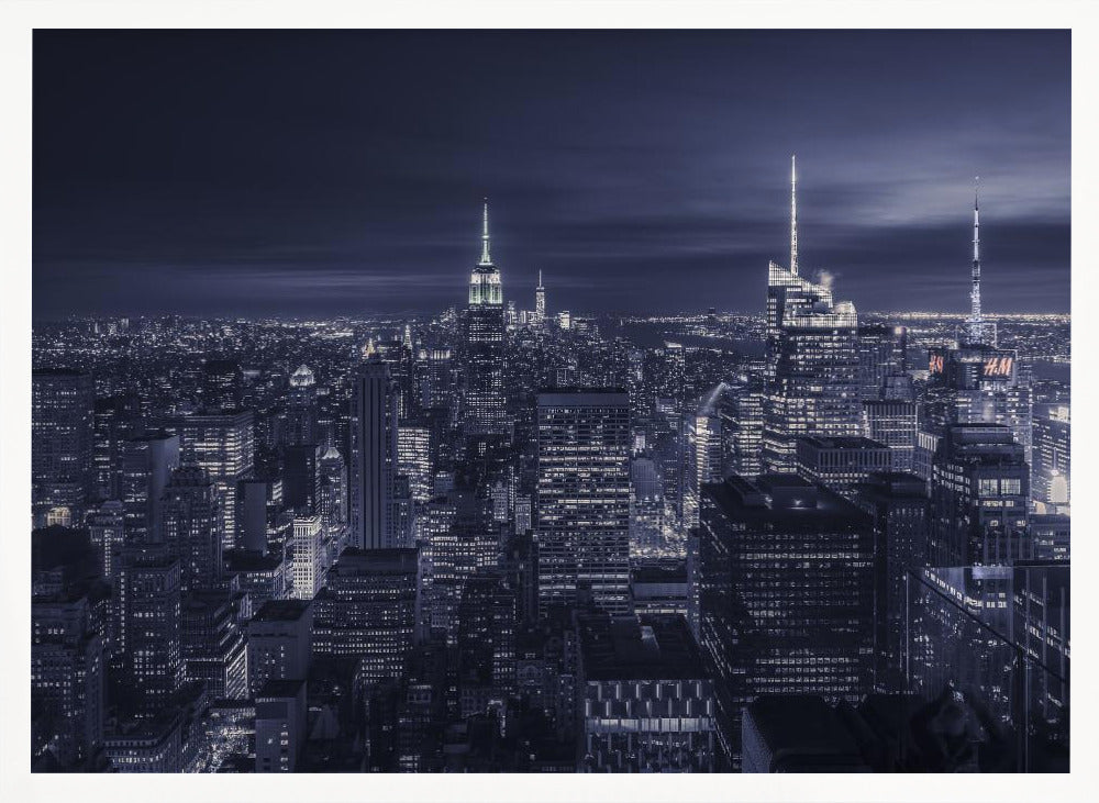 A dramatic nighttime photograph of the New York City skyline, viewed from a high vantage point. The cityscape is a sea of illuminated skyscrapers under a dark, moody sky, with the entire image cast in a cool blue and white monochrome tone and presented in a silver frame. Artwork