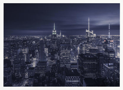 A dramatic nighttime photograph of the New York City skyline, viewed from a high vantage point. The cityscape is a sea of illuminated skyscrapers under a dark, moody sky, with the entire image cast in a cool blue and white monochrome tone and presented in a silver frame. Artwork