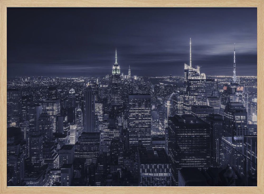A dramatic nighttime photograph of the New York City skyline, viewed from a high vantage point. The cityscape is a sea of illuminated skyscrapers under a dark, moody sky, with the entire image cast in a cool blue and white monochrome tone and presented in a silver frame. Artwork
