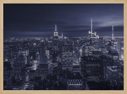 A dramatic nighttime photograph of the New York City skyline, viewed from a high vantage point. The cityscape is a sea of illuminated skyscrapers under a dark, moody sky, with the entire image cast in a cool blue and white monochrome tone and presented in a silver frame. Artwork