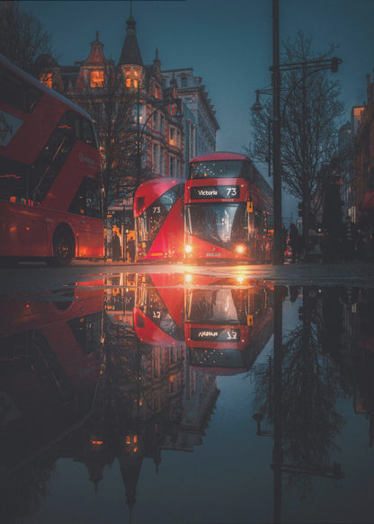 A moody, low-angle photograph of a wet London street at dusk, with a vibrant red double-decker bus reflected perfectly in a large puddle in the foreground. Print