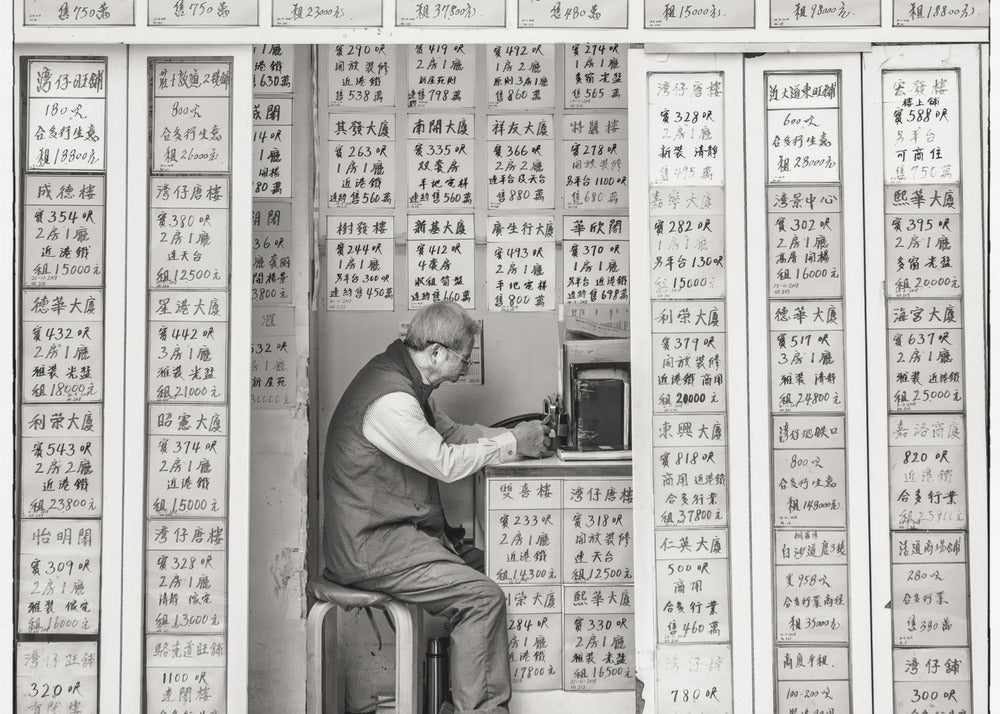 A black and white photograph of an elderly man with glasses sitting on a stool in a small, cramped shop. He is hunched over a counter, operating a small machine. The walls around him are completely covered with vertical paper signs featuring handwritten Chinese calligraphy, likely property listings or advertisements. Artwork