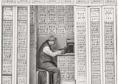 A black and white photograph of an elderly man with glasses sitting on a stool in a small, cramped shop. He is hunched over a counter, operating a small machine. The walls around him are completely covered with vertical paper signs featuring handwritten Chinese calligraphy, likely property listings or advertisements. Artwork