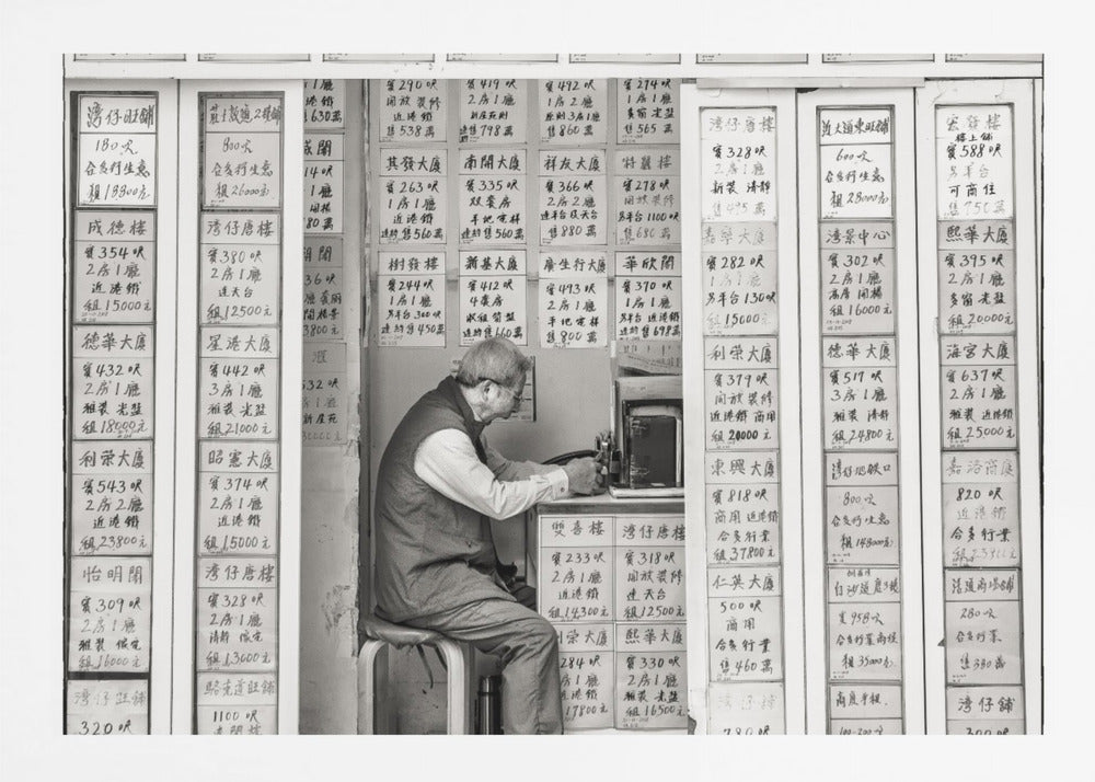 A black and white photograph of an elderly man with glasses sitting on a stool in a small, cramped shop. He is hunched over a counter, operating a small machine. The walls around him are completely covered with vertical paper signs featuring handwritten Chinese calligraphy, likely property listings or advertisements. Artwork