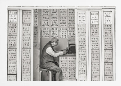 A black and white photograph of an elderly man with glasses sitting on a stool in a small, cramped shop. He is hunched over a counter, operating a small machine. The walls around him are completely covered with vertical paper signs featuring handwritten Chinese calligraphy, likely property listings or advertisements. Artwork