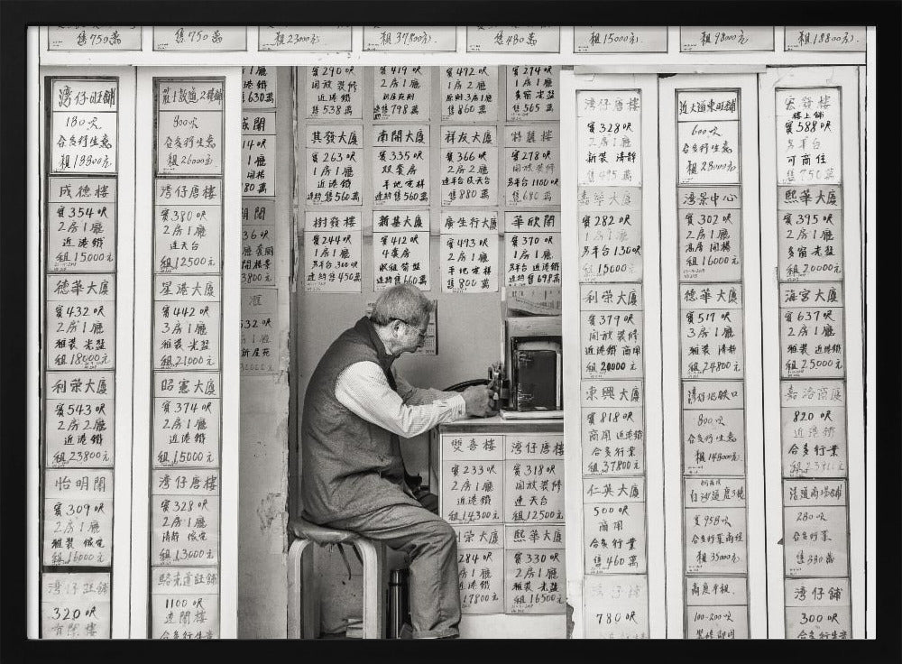 A black and white photograph of an elderly man with glasses sitting on a stool in a small, cramped shop. He is hunched over a counter, operating a small machine. The walls around him are completely covered with vertical paper signs featuring handwritten Chinese calligraphy, likely property listings or advertisements. Artwork