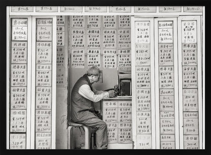A black and white photograph of an elderly man with glasses sitting on a stool in a small, cramped shop. He is hunched over a counter, operating a small machine. The walls around him are completely covered with vertical paper signs featuring handwritten Chinese calligraphy, likely property listings or advertisements. Artwork