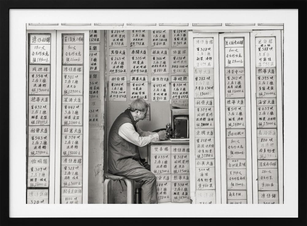 A black and white photograph of an elderly man with glasses sitting on a stool in a small, cramped shop. He is hunched over a counter, operating a small machine. The walls around him are completely covered with vertical paper signs featuring handwritten Chinese calligraphy, likely property listings or advertisements. Artwork