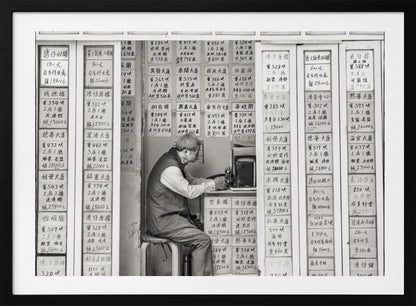 A black and white photograph of an elderly man with glasses sitting on a stool in a small, cramped shop. He is hunched over a counter, operating a small machine. The walls around him are completely covered with vertical paper signs featuring handwritten Chinese calligraphy, likely property listings or advertisements. Artwork