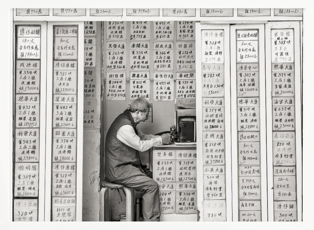A black and white photograph of an elderly man with glasses sitting on a stool in a small, cramped shop. He is hunched over a counter, operating a small machine. The walls around him are completely covered with vertical paper signs featuring handwritten Chinese calligraphy, likely property listings or advertisements. Artwork