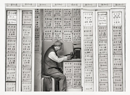 A black and white photograph of an elderly man with glasses sitting on a stool in a small, cramped shop. He is hunched over a counter, operating a small machine. The walls around him are completely covered with vertical paper signs featuring handwritten Chinese calligraphy, likely property listings or advertisements. Artwork
