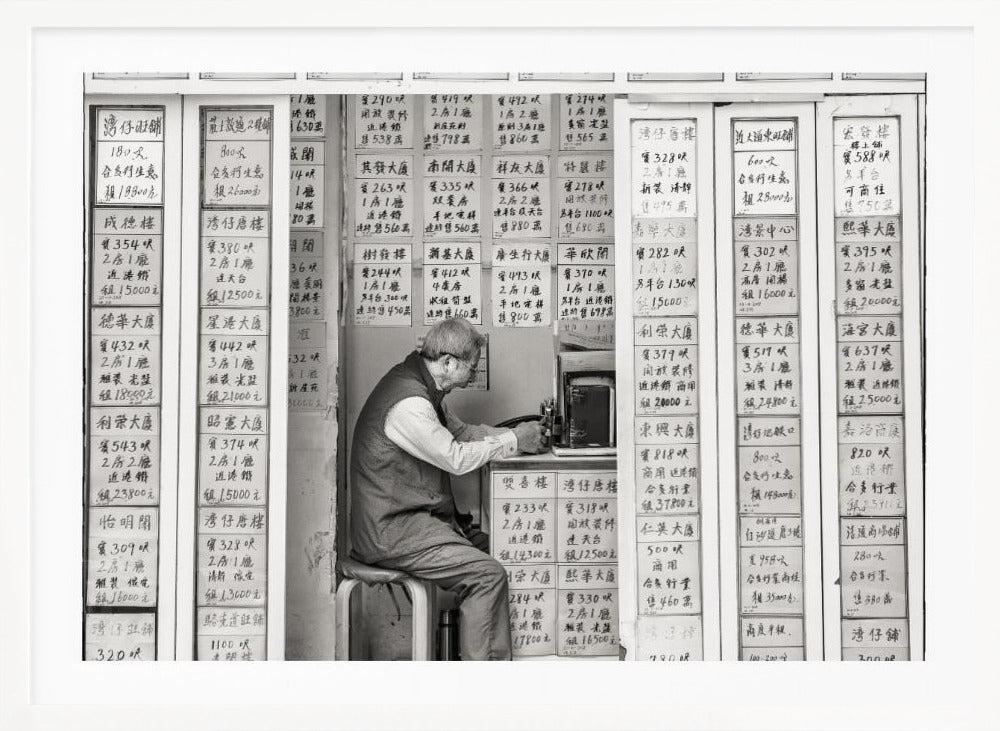 A black and white photograph of an elderly man with glasses sitting on a stool in a small, cramped shop. He is hunched over a counter, operating a small machine. The walls around him are completely covered with vertical paper signs featuring handwritten Chinese calligraphy, likely property listings or advertisements. Artwork