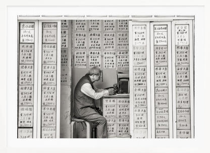 A black and white photograph of an elderly man with glasses sitting on a stool in a small, cramped shop. He is hunched over a counter, operating a small machine. The walls around him are completely covered with vertical paper signs featuring handwritten Chinese calligraphy, likely property listings or advertisements. Artwork