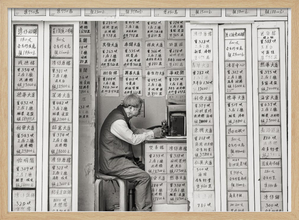 A black and white photograph of an elderly man with glasses sitting on a stool in a small, cramped shop. He is hunched over a counter, operating a small machine. The walls around him are completely covered with vertical paper signs featuring handwritten Chinese calligraphy, likely property listings or advertisements. Artwork