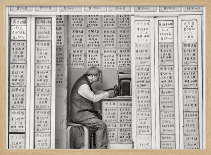 A black and white photograph of an elderly man with glasses sitting on a stool in a small, cramped shop. He is hunched over a counter, operating a small machine. The walls around him are completely covered with vertical paper signs featuring handwritten Chinese calligraphy, likely property listings or advertisements. Artwork