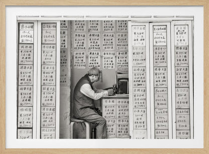 A black and white photograph of an elderly man with glasses sitting on a stool in a small, cramped shop. He is hunched over a counter, operating a small machine. The walls around him are completely covered with vertical paper signs featuring handwritten Chinese calligraphy, likely property listings or advertisements. Artwork