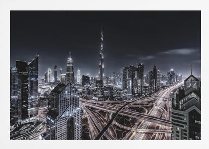 A striking black and white aerial photograph of the Dubai skyline at night, captured in a silver frame. The Burj Khalifa stands tall in the background, while a complex web of illuminated highway overpasses and light trails from traffic fills the foreground, creating a dynamic and futuristic cityscape. Print