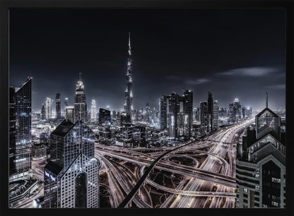A striking black and white aerial photograph of the Dubai skyline at night, captured in a silver frame. The Burj Khalifa stands tall in the background, while a complex web of illuminated highway overpasses and light trails from traffic fills the foreground, creating a dynamic and futuristic cityscape. Print
