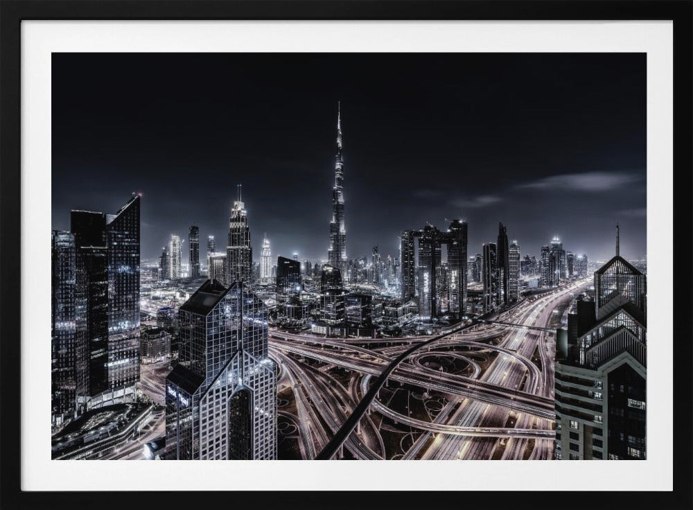A striking black and white aerial photograph of the Dubai skyline at night, captured in a silver frame. The Burj Khalifa stands tall in the background, while a complex web of illuminated highway overpasses and light trails from traffic fills the foreground, creating a dynamic and futuristic cityscape. Print