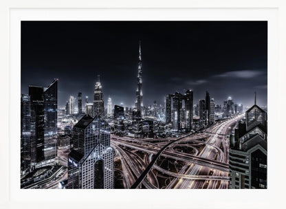 A striking black and white aerial photograph of the Dubai skyline at night, captured in a silver frame. The Burj Khalifa stands tall in the background, while a complex web of illuminated highway overpasses and light trails from traffic fills the foreground, creating a dynamic and futuristic cityscape. Print
