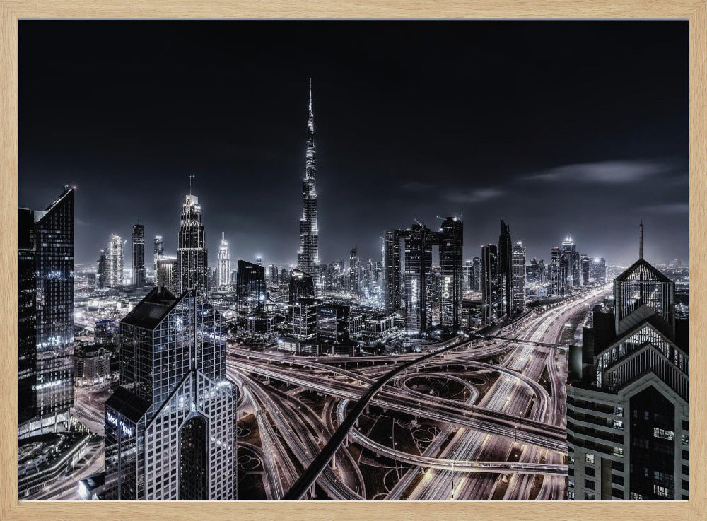 A striking black and white aerial photograph of the Dubai skyline at night, captured in a silver frame. The Burj Khalifa stands tall in the background, while a complex web of illuminated highway overpasses and light trails from traffic fills the foreground, creating a dynamic and futuristic cityscape. Print