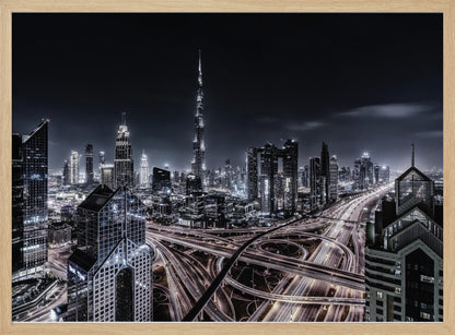A striking black and white aerial photograph of the Dubai skyline at night, captured in a silver frame. The Burj Khalifa stands tall in the background, while a complex web of illuminated highway overpasses and light trails from traffic fills the foreground, creating a dynamic and futuristic cityscape. Print