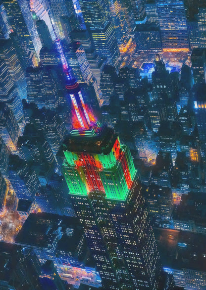 A dramatic high-angle aerial view of the Empire State Building at night, illuminated in festive red and green lights. The surrounding New York City skyline is a dense grid of skyscrapers glowing with blue and white lights, creating a vibrant, sprawling cityscape. Poster