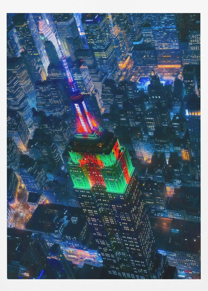 A dramatic high-angle aerial view of the Empire State Building at night, illuminated in festive red and green lights. The surrounding New York City skyline is a dense grid of skyscrapers glowing with blue and white lights, creating a vibrant, sprawling cityscape. Poster