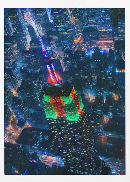 A dramatic high-angle aerial view of the Empire State Building at night, illuminated in festive red and green lights. The surrounding New York City skyline is a dense grid of skyscrapers glowing with blue and white lights, creating a vibrant, sprawling cityscape. Poster