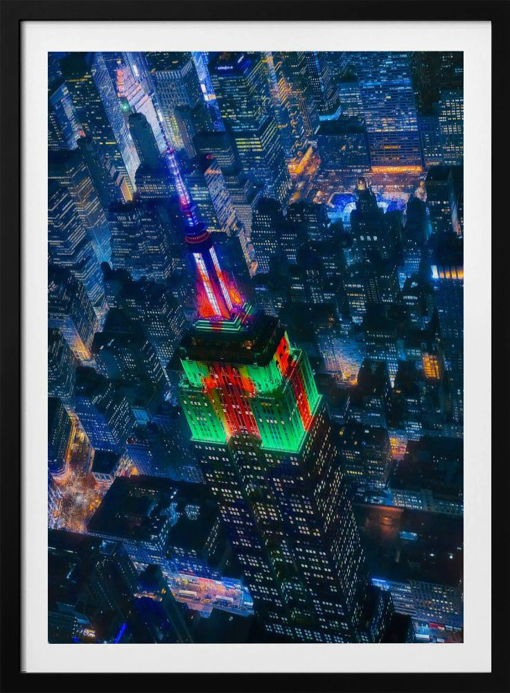A dramatic high-angle aerial view of the Empire State Building at night, illuminated in festive red and green lights. The surrounding New York City skyline is a dense grid of skyscrapers glowing with blue and white lights, creating a vibrant, sprawling cityscape. Poster