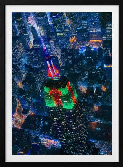A dramatic high-angle aerial view of the Empire State Building at night, illuminated in festive red and green lights. The surrounding New York City skyline is a dense grid of skyscrapers glowing with blue and white lights, creating a vibrant, sprawling cityscape. Poster