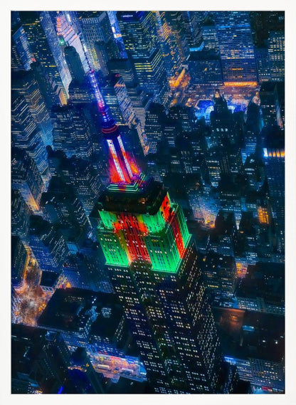 A dramatic high-angle aerial view of the Empire State Building at night, illuminated in festive red and green lights. The surrounding New York City skyline is a dense grid of skyscrapers glowing with blue and white lights, creating a vibrant, sprawling cityscape. Poster
