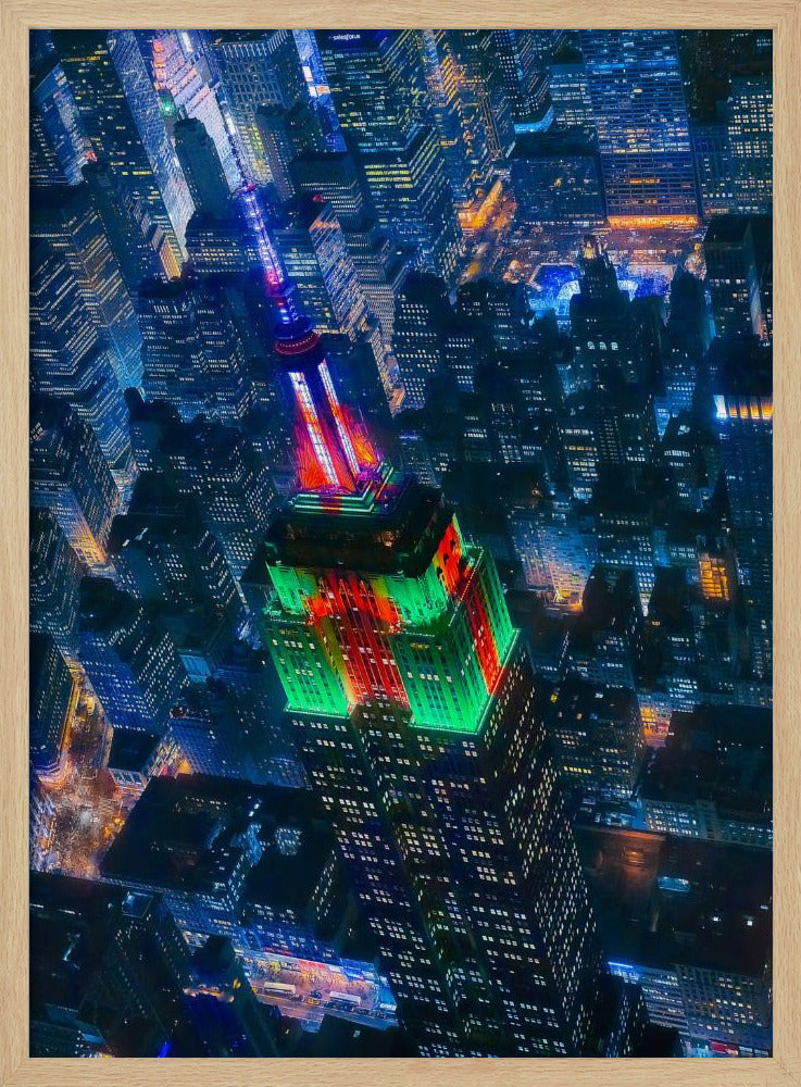 A dramatic high-angle aerial view of the Empire State Building at night, illuminated in festive red and green lights. The surrounding New York City skyline is a dense grid of skyscrapers glowing with blue and white lights, creating a vibrant, sprawling cityscape. Poster