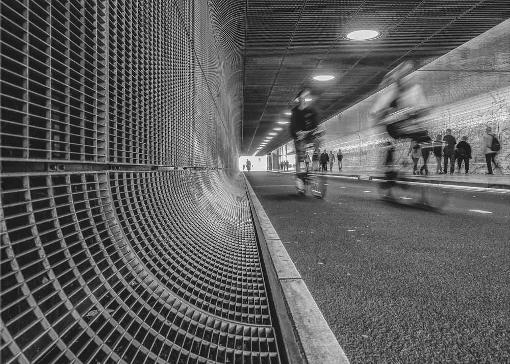 A dynamic black and white photograph from inside a modern tunnel. On the left, a curved metal grate wall creates a strong pattern, while on the right, cyclists are captured in motion blur on a paved path. Pedestrians are visible in the background, walking through the illuminated passage. Poster