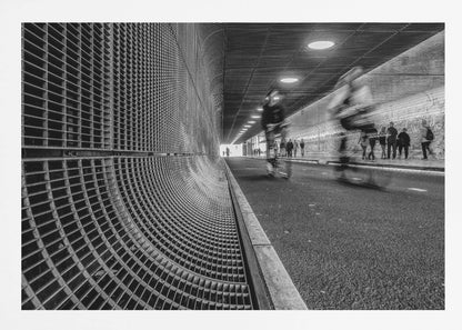 A dynamic black and white photograph from inside a modern tunnel. On the left, a curved metal grate wall creates a strong pattern, while on the right, cyclists are captured in motion blur on a paved path. Pedestrians are visible in the background, walking through the illuminated passage. Poster