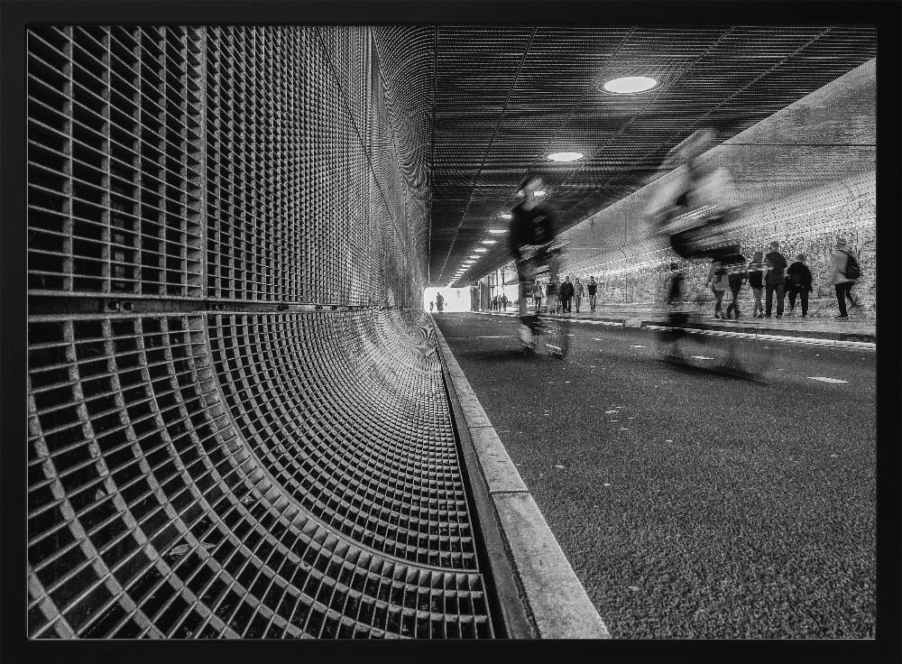 A dynamic black and white photograph from inside a modern tunnel. On the left, a curved metal grate wall creates a strong pattern, while on the right, cyclists are captured in motion blur on a paved path. Pedestrians are visible in the background, walking through the illuminated passage. Poster