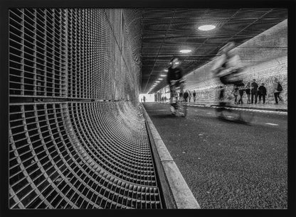 A dynamic black and white photograph from inside a modern tunnel. On the left, a curved metal grate wall creates a strong pattern, while on the right, cyclists are captured in motion blur on a paved path. Pedestrians are visible in the background, walking through the illuminated passage. Poster