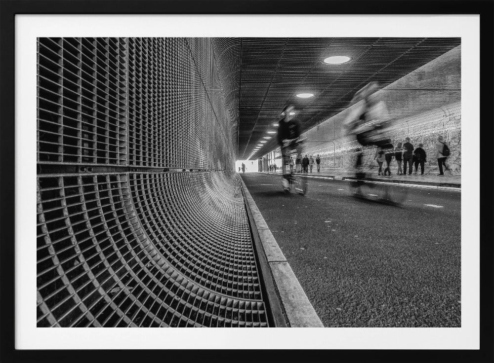 A dynamic black and white photograph from inside a modern tunnel. On the left, a curved metal grate wall creates a strong pattern, while on the right, cyclists are captured in motion blur on a paved path. Pedestrians are visible in the background, walking through the illuminated passage. Poster