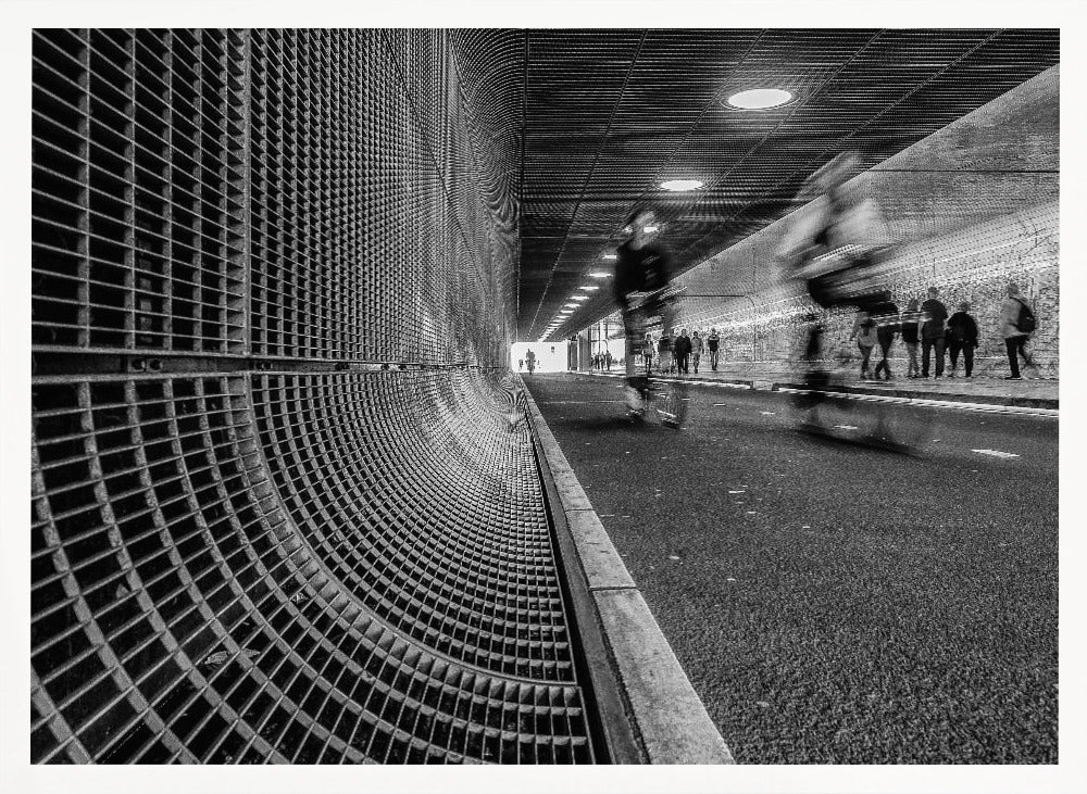A dynamic black and white photograph from inside a modern tunnel. On the left, a curved metal grate wall creates a strong pattern, while on the right, cyclists are captured in motion blur on a paved path. Pedestrians are visible in the background, walking through the illuminated passage. Poster