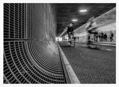 A dynamic black and white photograph from inside a modern tunnel. On the left, a curved metal grate wall creates a strong pattern, while on the right, cyclists are captured in motion blur on a paved path. Pedestrians are visible in the background, walking through the illuminated passage. Poster