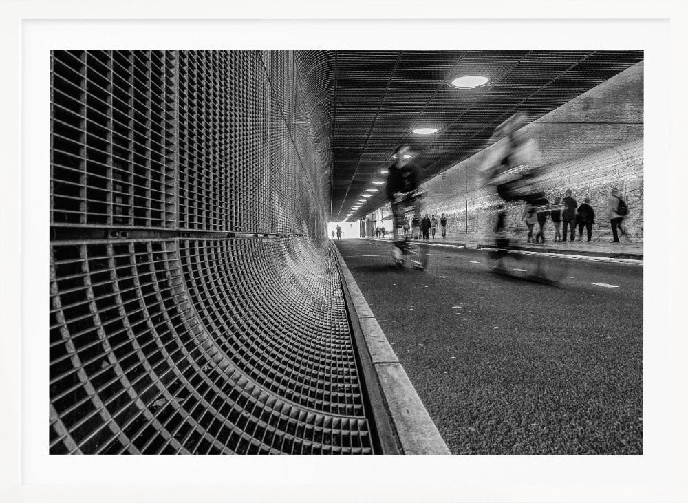 A dynamic black and white photograph from inside a modern tunnel. On the left, a curved metal grate wall creates a strong pattern, while on the right, cyclists are captured in motion blur on a paved path. Pedestrians are visible in the background, walking through the illuminated passage. Poster