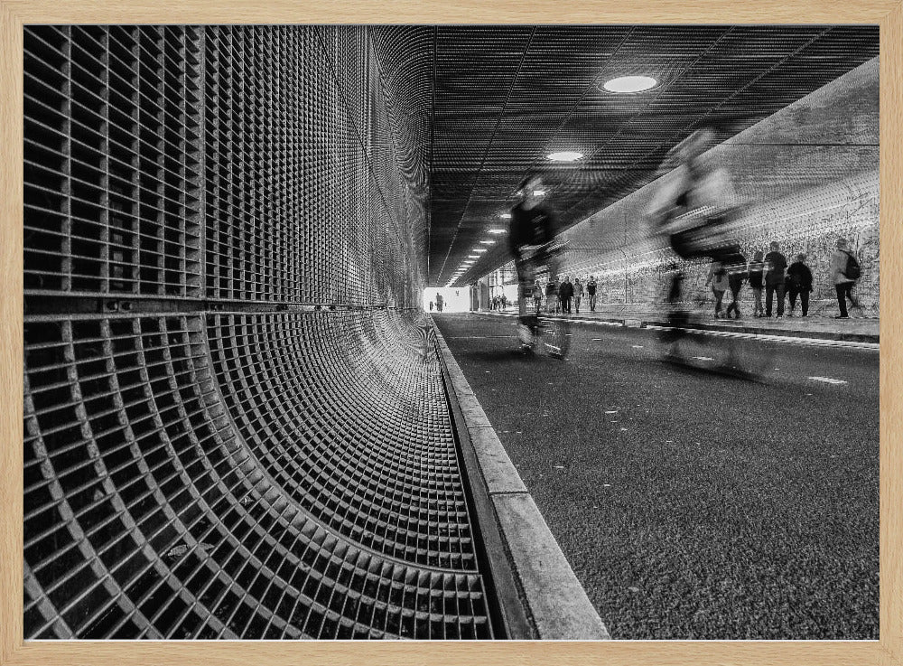 A dynamic black and white photograph from inside a modern tunnel. On the left, a curved metal grate wall creates a strong pattern, while on the right, cyclists are captured in motion blur on a paved path. Pedestrians are visible in the background, walking through the illuminated passage. Poster