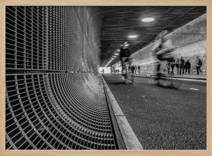 A dynamic black and white photograph from inside a modern tunnel. On the left, a curved metal grate wall creates a strong pattern, while on the right, cyclists are captured in motion blur on a paved path. Pedestrians are visible in the background, walking through the illuminated passage. Poster