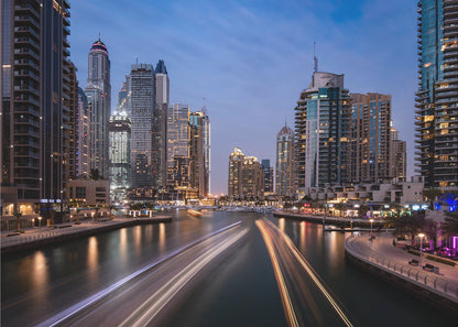 A long-exposure photograph of the Dubai Marina at dusk, framed in silver. Illuminated skyscrapers line a busy waterway, with light trails from moving boats creating long, bright streaks on the water. The sky is a deep twilight blue, and the city's lights cast a warm glow. Decor
