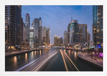 A long-exposure photograph of the Dubai Marina at dusk, framed in silver. Illuminated skyscrapers line a busy waterway, with light trails from moving boats creating long, bright streaks on the water. The sky is a deep twilight blue, and the city's lights cast a warm glow. Decor